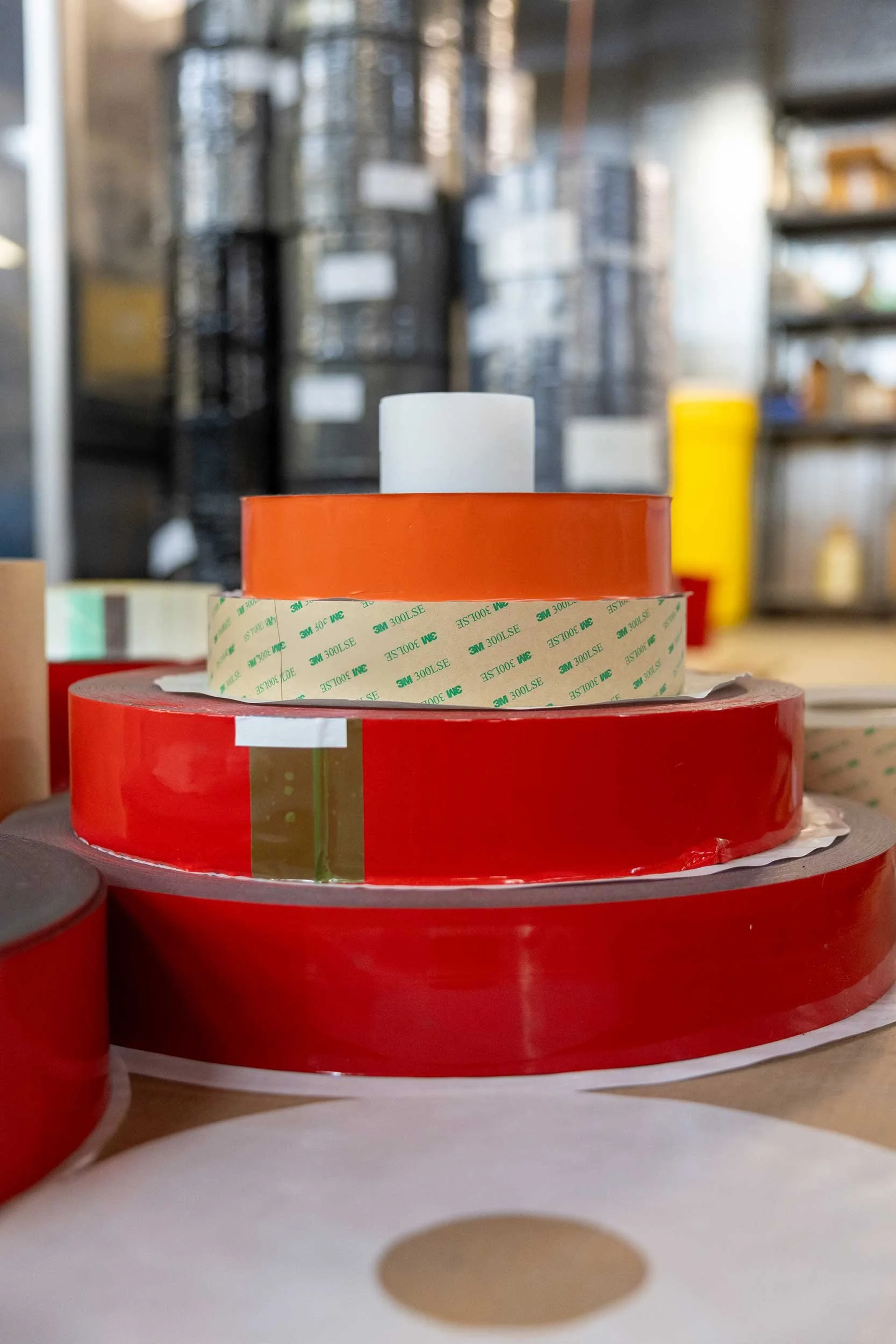 Large rolls of red, orange, and beige adhesive tape stacked on a table, with a blurred industrial warehouse background.
