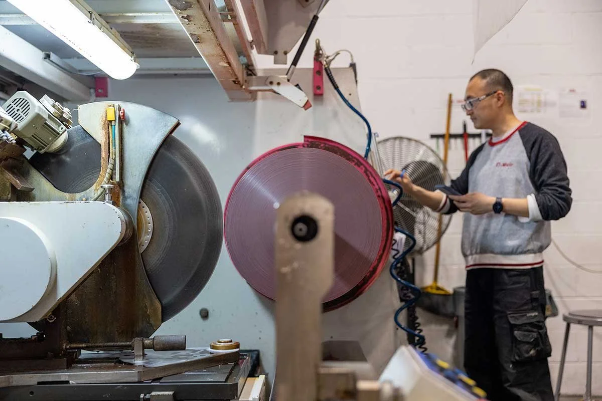 A man wearing glasses and a gray and black shirt appears to be operating a large industrial saw or cutting machine in a workshop or manufacturing setting. He is holding a clipboard or tablet and is focusing on the machine, which has a large spinning circular blade.