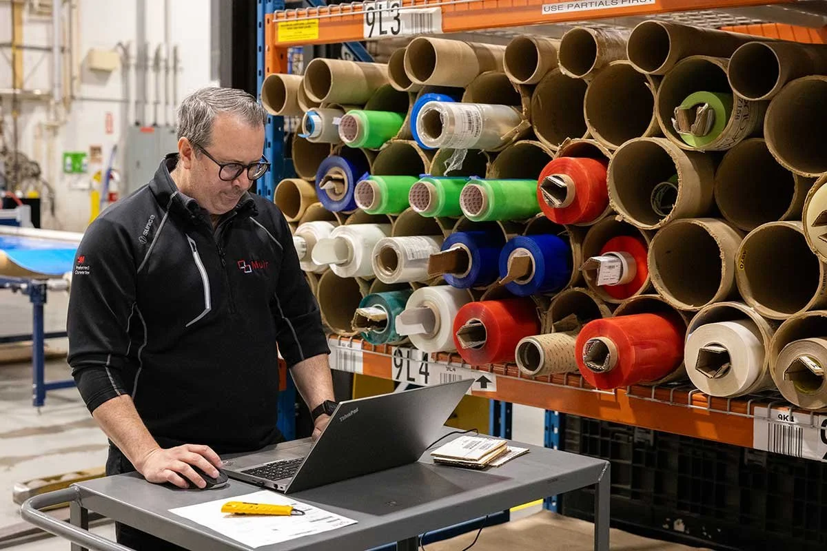 A man with glasses wearing a black jacket with a company logo standing at a table with a laptop, in an industrial warehouse, surrounded by shelves filled with large rolls of colored paper or vinyl.