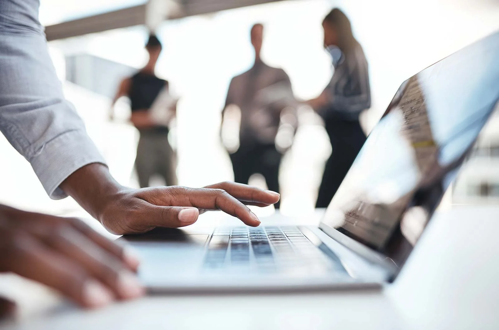 Close-up of a person's hand typing on a laptop keyboard with three blurred individuals standing in the background.