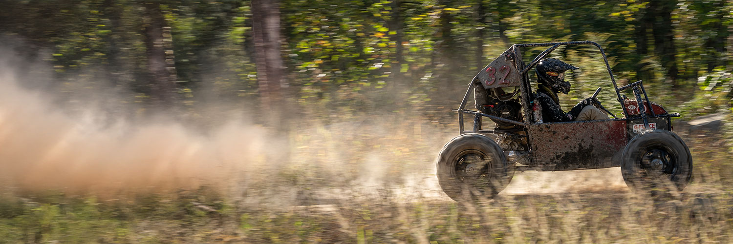 An off-road vehicle speeds through a dusty forest trail, kicking up dirt. Tall trees with green leaves line the path, and the motion blur emphasizes the vehicle's fast movement.